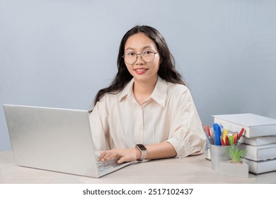 young woman is sitting at a desk working with her laptop. She wears glasses and a light-colored blouse. She smiles as she works at her laptop. On the desk, there are books stacked on one side  - Powered by Shutterstock