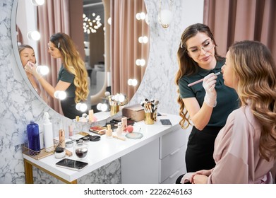 Young woman sitting at dressing table and looking in the mirror while makeup artist applying concealer with cosmetic brush. Female stylist doing professional makeup for client in beauty salon. - Powered by Shutterstock