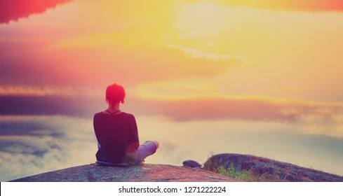 Young woman sitting enjoying peaceful moment of beautiful colorful sunset. In the reflection of the lake water sees clouds and sun. Vintage mood, concepts of winner, freedom, happiness.