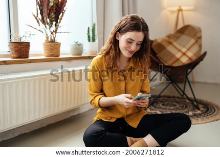 Young woman sitting in front of a radiator cross-legged on the floor with her mobile phone smiling to herself as she reads a text message