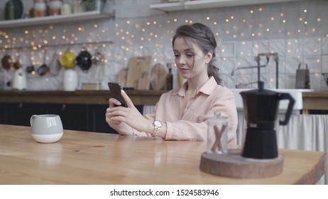 Young Woman Sitting At Her Kitchen Table At Home Working On Her Small Business With A Smart Phone. Stock Footage. Portrait Of A Young Lady Tapping Oh Her Mobile Phone.