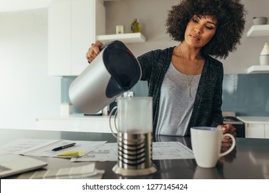 Young Woman Sitting At Her Working Table Pouring Water In A Jug. African Female Making Coffee For Her Self While Working At Home Office.