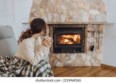 Young woman sitting at home by the fireplace with a hot tea or coffee mug and warming her hands, she is wearing white woollen sweater. Cold houses in Europe concept during energy and gas crisis.