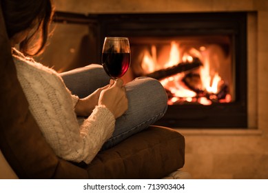 Young woman sitting at home by the fireplace and drinking a red wine.