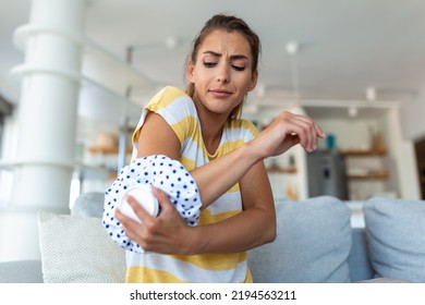 Young Woman Is Sitting At Home On Her Sofa And Touching The Her Elbow While Suffering From Elbow Pain, Placing Ice Pack On Hurt Spot