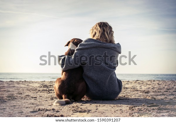 Young woman sitting and hugging\
dog on the beach. Friendship concept - woman and dog sitting\
together on a beach and enjoying sunrise. Back view colorized\
image