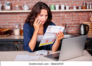 Young woman sitting in the kitchen with dissatisfied face in front of laptop holding utility bills, talking on mobile phone 