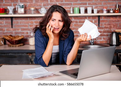 Young woman sitting in the kitchen with dissatisfied look on her face in front of laptop holding utility bills, talking on mobile phone 