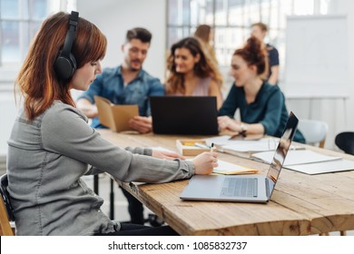 Young woman sitting listening to dictation or internet audio on headphones in a busy modern open plan office with her young colleagues working in the background - Powered by Shutterstock