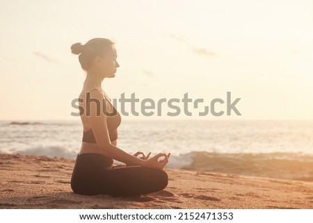 Young woman sitting in lotus position and does yoga for healthy lifestyle on ocean beach. Female performing sports exercises to restore strength and spirit. Yoga position on seashore at sunset sea