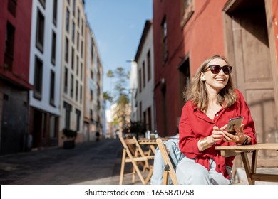 Young Woman Sitting With Mobile Phone On The Cafe Terrace On The Old City Steet During A Sunny Day. Street View On The Old Spanish Town La Laguna