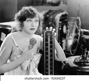 Young woman sitting next to a fan and a thermometer looking hot and eating an ice cream
