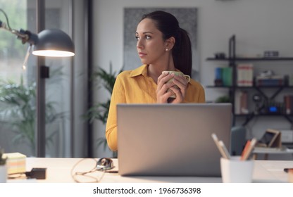 Young Woman Sitting At Office Desk And Drinking Coffee, She Is Looking Outside The Window