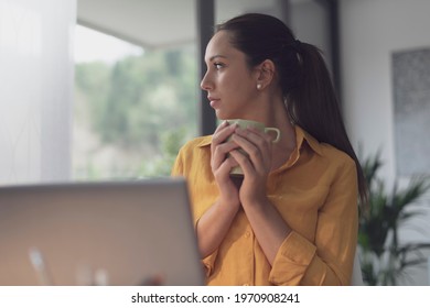 Young Woman Sitting At Office Desk And Drinking Coffee, She Is Looking Outside The Window