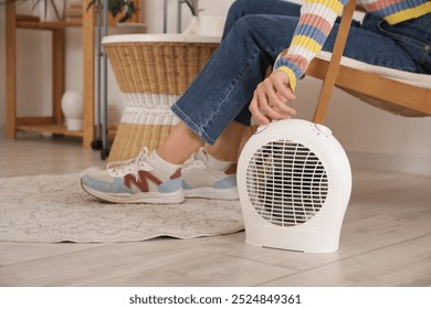 Young woman sitting on armchair near electric fan heater in living room, closeup