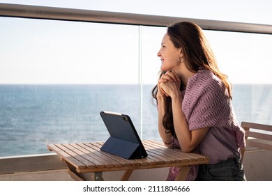 A young woman is sitting on the balcony with a tablet and looking at the seashore