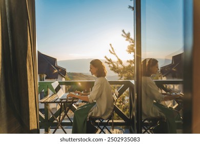 Young woman sitting on balcony with view of mountains and sunset working using laptop - Powered by Shutterstock