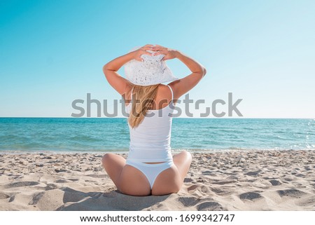 Young woman sitting on a beach holding her straw hat and looking to the horizon.
