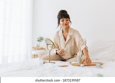 Young woman sitting on bed at home and applying cosmetics. Woman using skin care products at home. - Powered by Shutterstock