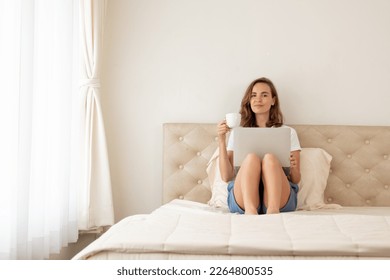 Young woman sitting on bed and using laptop. Beautiful Young woman using laptop on bed at home in the morning. Young female using laptop, sitting on bed in hotel. - Powered by Shutterstock