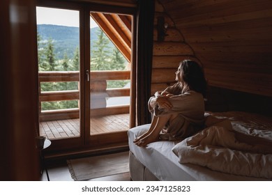 Young woman sitting on a bed in a wooden chalet cabin in the mountains. Comfortable rest and sleep in a log house in nature - Shutterstock ID 2347125823