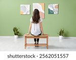 Young woman sitting on bench near paintings in art gallery, back view