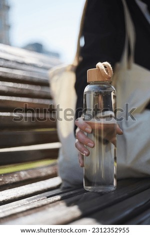 Young woman sitting on a bench in the morning and holding a glass water bottle in hand. Unrecognizable female person chilling outdoor in a sunny day with an eco bottle of fresh water