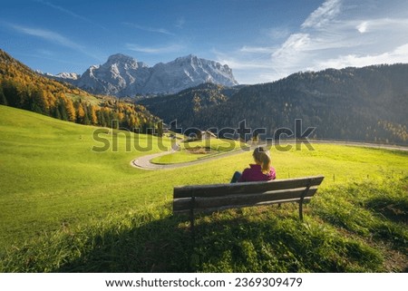 Young woman sitting on the bench and beautiful alpine village at sunset in autumn. Tyrol, Dolomites, Italy. Colorful landscape with girl, green meadows, orange trees, road, mountain, blue sky in fall