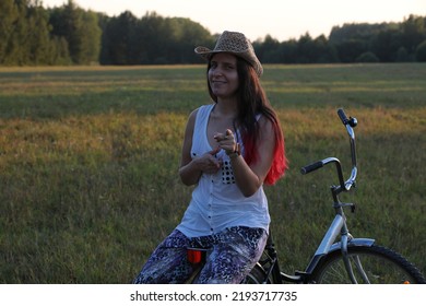 A Young Woman Is Sitting On A Bicycle In A Field In Nature Looking At The Camera With A Sly Smile On Her Face And Jokingly Pointing Her Finger