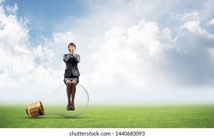 Young Woman Sitting On Big Light Bulb. Girl In Business Suit Keeps Hands On Face For Surprise. Businesswoman Playing Hide And Seek On Green Meadow. Hide From Problems. Help And Successful Solutions