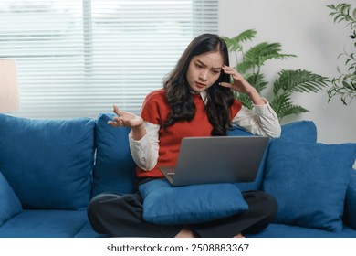 Young woman is sitting on a blue couch and looking stressed while working from home on her laptop - Powered by Shutterstock