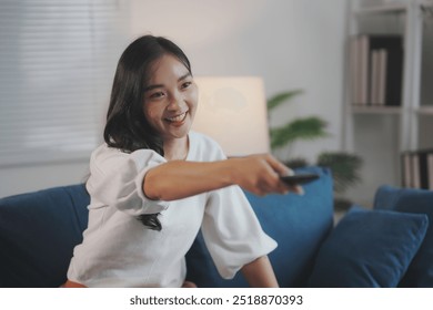 Young woman is sitting on a blue sofa in her living room, smiling and using a remote control to change channels on the television - Powered by Shutterstock