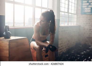 Young Woman Sitting On A Box At Gym After Her Workout. Female Athlete Taking Rest After Exercising At Gym.