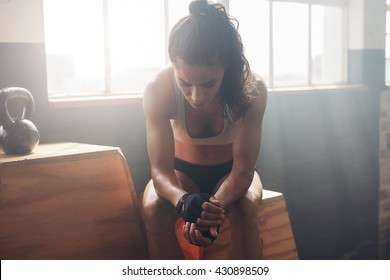 Young Woman Sitting On A Box At Gym After Her Workout And Looking Down. Female Athlete Taking Rest After Fitness Training At Gym.