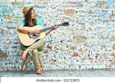 Young woman sitting on chair play guitar. Vintage brick wall background.