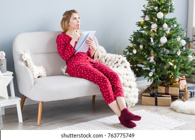 Young Woman Is Sitting On A Comfortable Sofa In  Pajamas At Home And Making Plans For Christmas And New Year Celebration. A Girl Is Thinking With A Notepad And Pen In Hands