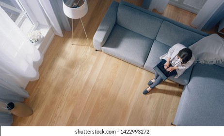 Young Woman Is Sitting On A Couch, Working Or Studying On A Laptop. Cozy Living Room With Modern Interior, Grey Sofa And Wooden Flooring. Top View Camera Shot.