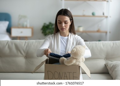 Young woman sitting on couch preparing parcel for sending to needy human. Girl with big kind heart puts used clothes new wear and soft bear toy in donation box, concept of caring about homeless people