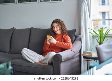 Young woman sitting on couch holding smartphone, looking at cellphone using cell phone checking mobile apps, reading messages, doing ecommerce shopping, chatting online, watching videos, playing game.