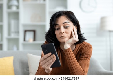 Young woman sitting on a couch at home, looking at her smartphone with a bored expression. Concept of boredom, technology addiction, and home lifestyle. - Powered by Shutterstock