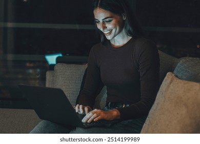 Young woman sitting on a couch in a hotel room, using a laptop in the evening. Engaged in remote work or chatting with friends and family, embracing the digital nomad lifestyle while traveling  - Powered by Shutterstock