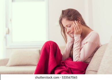 Young woman sitting on a couch, holding her head, having a strong headache. Lens flare effect on the window