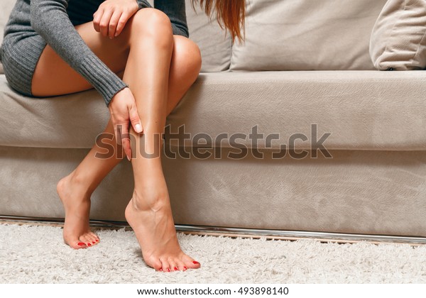 Young woman sitting on the couch suffering from\
severe pain in the leg