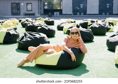 A Young Woman Is Sitting On An Easy Chair Outdoors And Enjoying A Sunny Day. A Beautiful Contented Girl In Sunglasses.