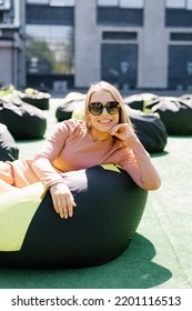 Young Woman Is Sitting On An Easy Chair Outdoors And Enjoying A Sunny Day. A Beautiful Contented Girl In Sunglasses.