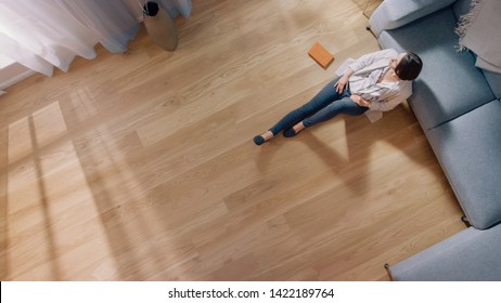 Young Woman Is Sitting On A Floor And Looking Out Of The Window After Reading A Book. Cozy Living Room With Modern Interior, Grey Sofa And Wooden Flooring. Top View Camera Shot.