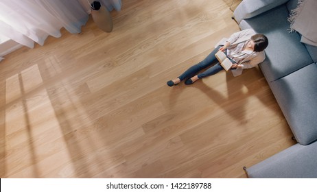 Young Woman Is Sitting On A Floor And Reading A Book. Cozy Living Room With Modern Interior, Grey Sofa And Wooden Flooring. Top View Camera Shot.