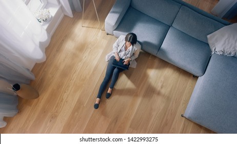 Young Woman Is Sitting On A Floor, Working Or Studying On A Laptop. Cozy Living Room With Modern Interior, Grey Sofa And Wooden Flooring. Top View Camera Shot.