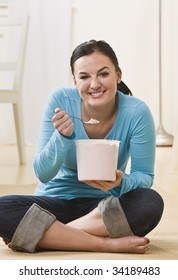 A Young Woman Is Sitting On The Floor And Eating Ice Cream Out Of A Tub.  She Is Smiling At The Camera.  Vertically Framed Shot.