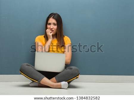 Young woman sitting on the floor with a laptop biting nails, nervous and very anxious